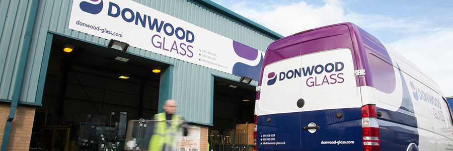 A warehouse with a Donwood Glass sign and a purple and white van. A person in a high-visibility vest walks in the foreground.